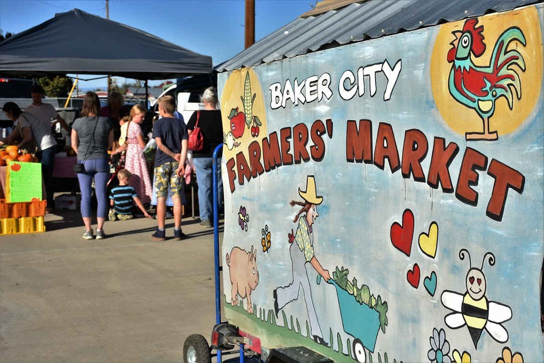 Ein farbenfrohes, handgemaltes Schild für einen Bauernmarkt mit Illustrationen landwirtschaftlicher Elemente wie einem Hahn, Mais und Äpfeln. Im Hintergrund sind Marktstände und Besucher zu sehen.