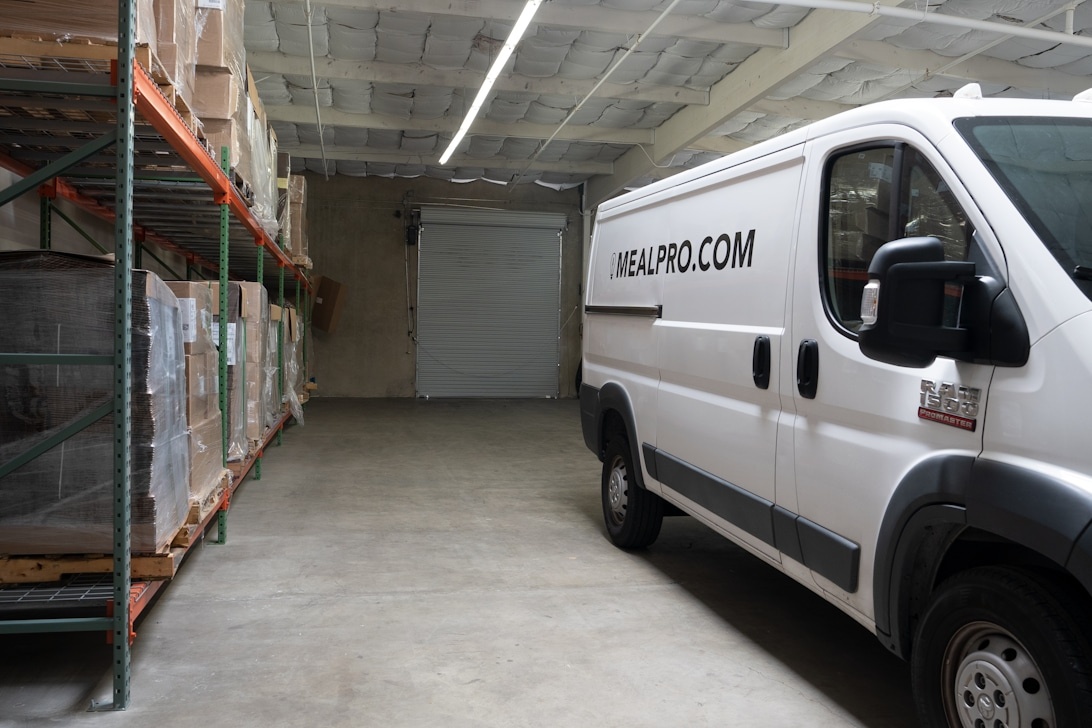 Ein Lieferwagen der Marke MEALPRO.COM in einer Lagerhalle mit sichtbaren Regalbeständen, was integrierte Kleinunternehmenslogistik veranschaulicht, die Lagerung, Fulfillment und Lieferdienste kombiniert.
